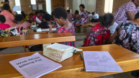 Course booklets and an open Bible sitting on a church bench looking into the church from the back, with lots of women studying and talking in small groups in the back ground