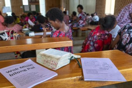 Course booklets and an open Bible sitting on a church bench looking into the church from the back, with lots of women studying and talking in small groups in the back ground