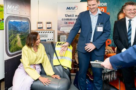 A woman sits on an aircraft seat whilst a man beckons another to sit