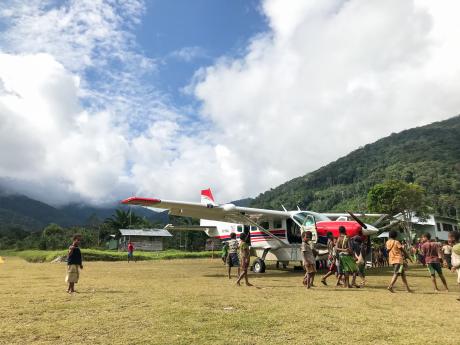 MAF aircraft parked at an remote, grassy airstrip with people all gathered around and crowding under its left wing